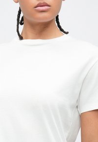 Άτομο με πλεκτή κότσο, φοράει απλό λευκό κοντομάνικο μπλουζάκι με στρογγυλή λαιμόκοψη, στο φόντο ανοιχτού χρώματος.