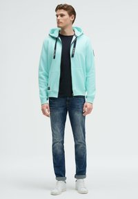 Felpa con zip verde menta dotata di cappuccio, dettagli neri e lacci, indossata sopra una maglietta nera; abbinata a jeans blu e sneakers bianche.