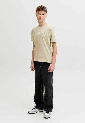 Teenager med krøllet hår iført beige t-shirt med grafik, sorte løse jeans og hvide sneakers, stående foran en ensfarvet hvid baggrund.
