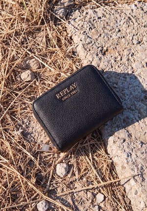 Una cartera negra de piel Replay con cremallera está sobre hierba seca y terreno rocoso.