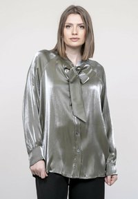 Blouse verte métallique avec un nœud au col, manches longues et fermeture à boutons sur le devant. Texture lisse avec une finition brillante.