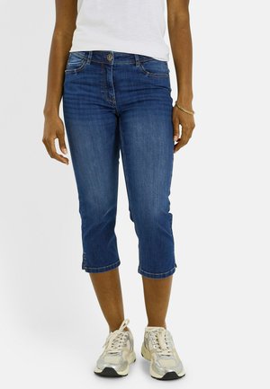 Persona con vaqueros cropped de tiro medio de color azul oscuro, zapatillas blancas, una camiseta blanca y una pulsera dorada en la muñeca derecha.