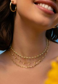 Colliers en couches dorées avec des perles vertes, une chaîne en forme de trombone et des accents pendants sur un fond de peau lisse. Boucles d'oreilles en forme de cerceau.