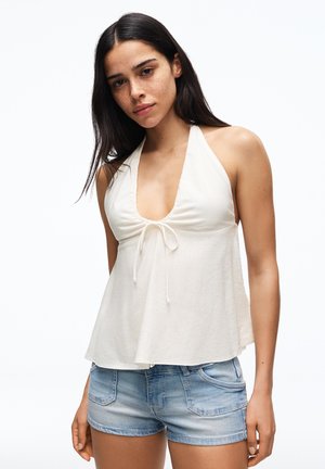 Žena s dlhými tmavými vlasmi, oblečená v bielom tielku bez rukávov s halter nákrčníkom a svetlomodrých džínsových šortkách, stojaca na bielom pozadí.