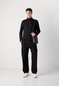 Svart langermet skjorte med høy krage og logo, kombinert med svarte cargo-bukser med sidelommer og hvite joggesko.