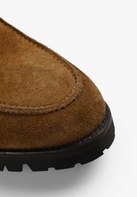 Brune ruskindsstøvle med rund tå, detaljeret syning og en tekstureret sort gummibund med dybe mønstre for godt greb.