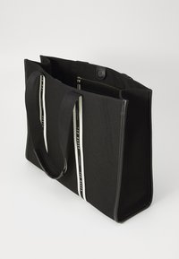 Schwarze Leinentasche mit zwei vertikalen weißen Streifen und dem Text "TED BAKER". Offene Oberseite mit magnetischem Verschluss, robuste Griffe und ein elegantes Design.