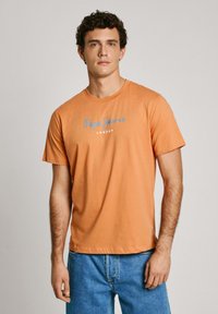 Oranžinė medvilninė T-shirt trumpomis rankovėmis, su mėlynu ir baltu "Pepe Jeans London" logotipu priekyje. Lygus paviršius ir standartinė forma.