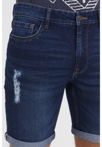 Tamno plave traper kratke hlače s poderanim dijelom sprijeda. Imaju uvrnute rubove i standardne džepove. Završetak s bakrenim detaljima.