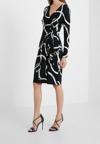 Svart omslagsklänning med långa ärmar som har ett vitt kedjemönster; knälång, mjukt tyg och en knytdetalj i midjan. Kombinerad med svarta högklackade sandaler.