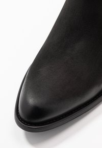 Bota de cuero negra hasta el tobillo con punta afilada, textura suave y un sutil brillo. Presenta una costura de contraste a lo largo de la suela.