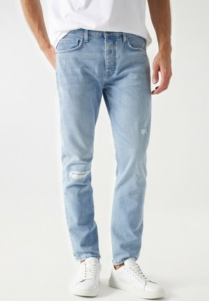 Homme portant un jean bleu clair usé, des baskets blanches et une chemise blanche sur un fond blanc uni.