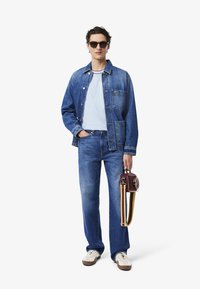 Giacca di denim e jeans di un blu medio, abbinati a una t-shirt azzurro chiaro. Tiene una piccola borsa bordeaux con tracolle gialle e rosse.