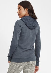 Haut à capuche bleu marine avec une capuche en tricot texturé à motif en cable, à manches longues et avec des poignets côtelés. Porté avec un pantalon à motif carreaux.