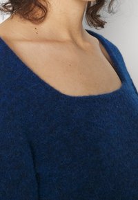 Blå ulltröja med en bred, fyrkantig halsringning och mjuk, fluffig textur. Tyget verkar tjockt och bekvämt, lämpligt för vardagsbruk.