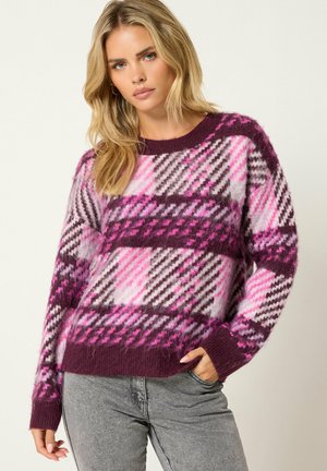 Femme aux longs cheveux blonds portant un pull à carreaux violet, rose et blanc et un jean gris, debout avec une main dans la poche.