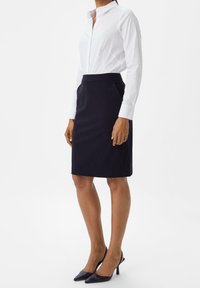 Weißes Hemd mit Knopfleiste und hohem Kragen, kombiniert mit einem taillierten dunkelblauen Rock und schwarzen spitzen Pumps, die einen eleganten, professionellen Look zeigen.