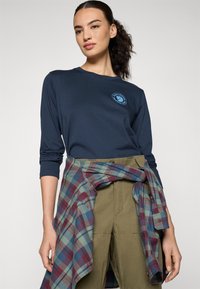 Marineblauwe lange mouwen shirt met blauw logo; groene broek met een flanellen shirt om de taille geknoopt, met een patroon van blauwe, rode en groene ruit.