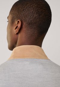 Suéter gris con un cuello de ante beige, con una marca en relieve. La tela es acanalada, mostrando un detalle texturizado en el cuello.