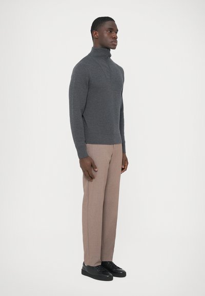 Camisola de gola alta cinzenta em malha com design de fecho de correr pela metade, combinada com calças bege e ténis pretos, mostrada de lado sobre um fundo neutro.