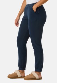 Personne portant un pantalon de jogging en polaire bleu marine et des chaussons beige à enfiler avec une doublure en fourrure blanche sur un fond uni.