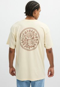 Camiseta beige de corte holgado, con un gran diseño floral marrón en la parte posterior, fabricada en suave tejido de algodón. Manga corta.