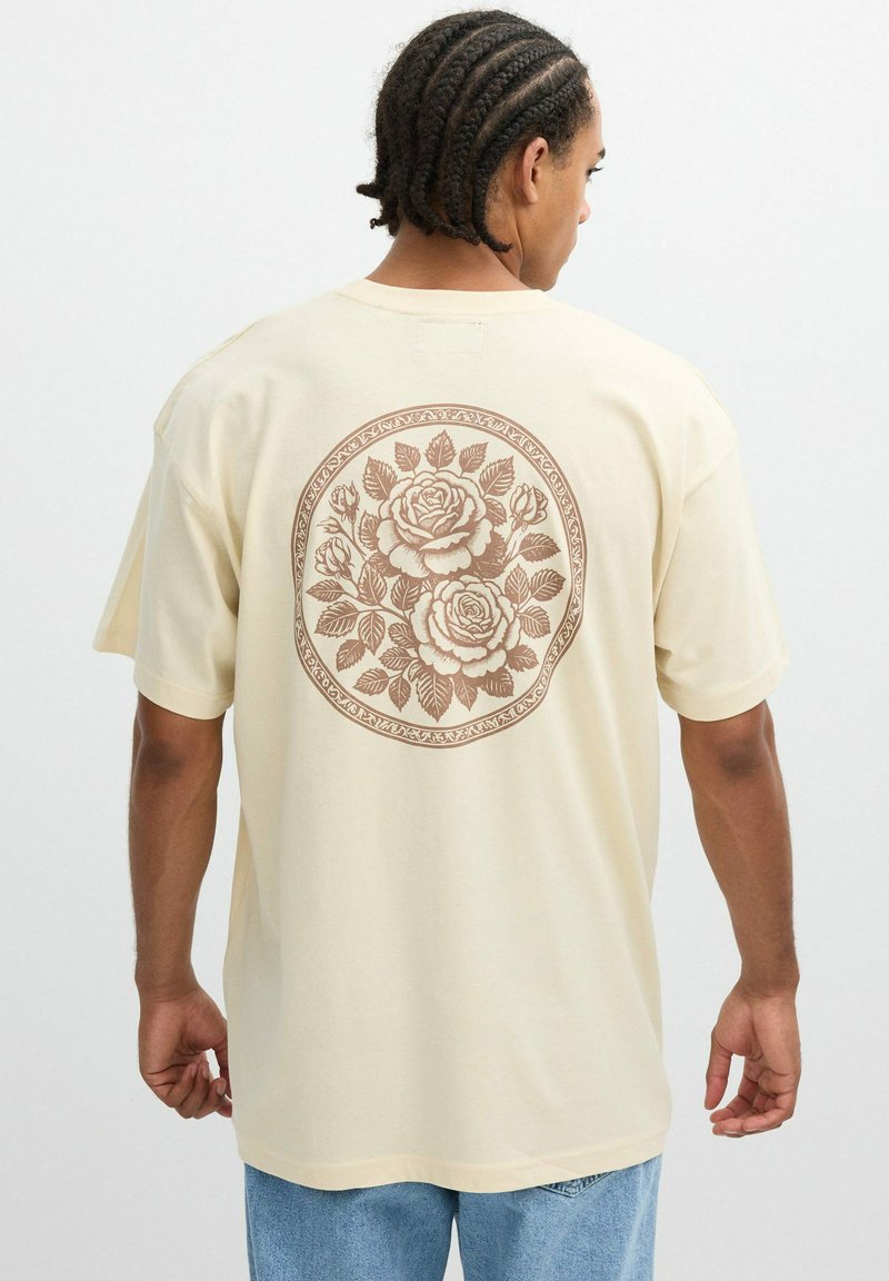 Camiseta beige de corte holgado, con un gran diseño floral marrón en la parte posterior, fabricada en suave tejido de algodón. Manga corta.