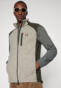 Homem em colete de fleece bege e verde, por cima de uma camisa de mangas longas cinza, calças de corduroy castanhas e óculos de sol beges, de pé com as mãos nos bolsos.