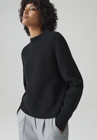 Maglione nero lavorato a maglia con un collo alto e maniche lunghe; con costine testurizzate; abbinato a pantaloni grigi chiari a pieghe.
