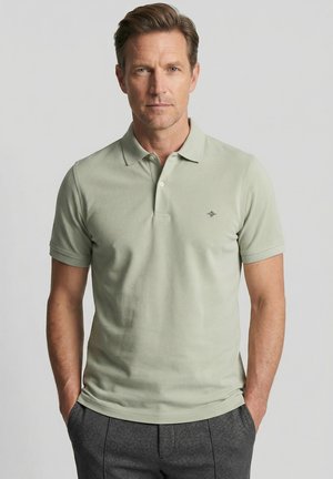 Man met lichtbruin haar, gekleed in een lichtgroene polo en grijze broek, staat met handen in de zakken tegen een effen achtergrond.