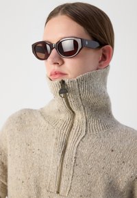 Pull de malha bege com gola alta e zíper de um quarto; apresenta um padrão texturizado e um pequeno logotipo redondo em metal. Óculos de sol geométricos castanhos.