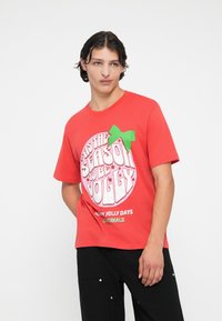 Tricou roșu din bumbac, cu un grafic circular alb care conține text și un accent de fundiță verde pe partea stângă. Pantaloni negri vizibili dedesubt.