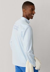 Homme souriant, portant une chemise à rayures bleu clair avec un texte en écriture au dos, un pantalon bleu, tenant un pull de couleur crème sur son bras.