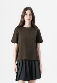 Brown cotton t-shirt with short sleeves and a rounded neckline, layered over a pleated black skirt. Simple, casual design with a relaxed fit.