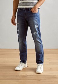 Vaqueros de mezclilla de color azul claro con detalles desgastados y parches rasgados. Diseño ajustado combinado con zapatillas blancas, contra un fondo neutro.