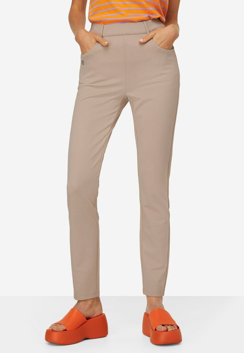 Béžové slim-fit kalhoty s předními kapsami; hladká textura; spárované s oranžovými platformovými sandály. Žádné viditelné vzory ani kovové prvky.