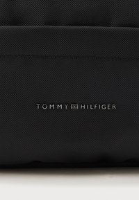 Čierny nylonový materiál s textúrovaným povrchom, s embosovaným logom "TOMMY HILFIGER" v striebornej farbe nad jednoduchým horizontálnym švom.