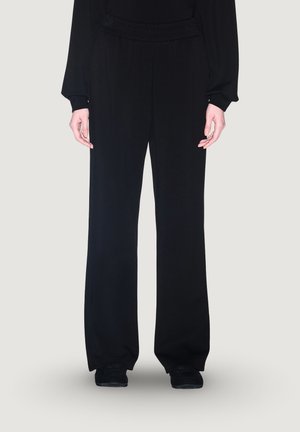 Partie inférieure du corps d'une personne portant un pantalon noir ample, des chaussures noires et un haut noir à manches longues sur un fond clair uni.