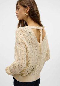 Maglione lavorato a maglia color beige chiaro con scollatura a V sul retro, dotato di un sottile nastro e motivo a traforo. Vestibilità ampia e orli a coste.