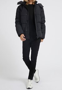 Sort dun jakke med lynlås og aftagelig hætte, med quiltet design og glat tekstur. Kombineret med sorte bukser og hvide sneakers.