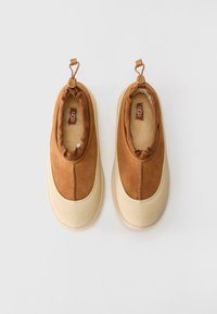 Pár hnědých semišových slip-on bot UGG s krémovými gumovými podrážkami a měkkým fleecovým podšívkou, zobrazených shora na bílém pozadí.