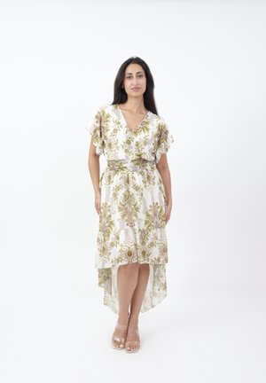 Kobieta stojąca w białej sukience z zielonymi i brązowymi motywami kwiatowymi, z falbaniastymi rękawami i asymetrycznym dołem, w połączeniu z szpilkami na przezroczystych paskach.