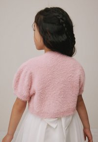 Cardigan roz cu mâneci scurte, pufoasă, cu o suprafață texturată, purtat peste o rochie albă cu o fundă în talie. Vedere din spate.