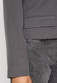 Blazer grigio in tessuto liscio, con una tasca applicata e maniche strutturate, abbinato a pantaloni grigi scuri con texture.