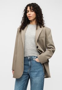 Μπεζ σακάκι με εφαρμογή, με δομημένους ώμους, κλείσιμο με ένα κουμπί και μία τσέπη στο στήθος, σε συνδυασμό με ανοιχτό γκρι πουλόβερ και μπλε τζιν.