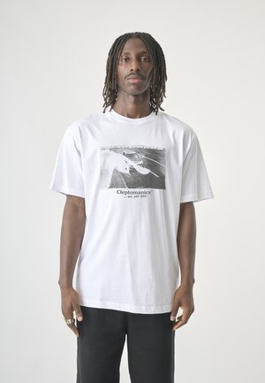 Man met dreadlocks die een wit grafisch T-shirt draagt met een zwart-wit foto en tekst met de tekst "Cleptomanicx ...we got you."