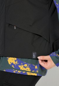 Schwarze Jacke mit farbenfrohem Spritzdesign, verfügt über geschichteten Stoff und ein Etikett mit der Aufschrift "HYPAR". Texturierte und glatte Bereiche sind sichtbar.