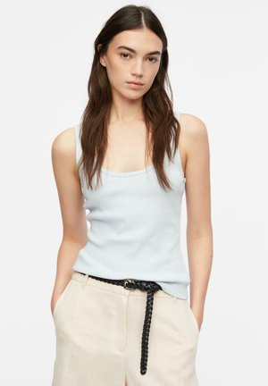 Młoda kobieta z długimi brązowymi włosami, ubrana w jasnoniebieską bezrękawnik, beżowe spodnie i czarny pleciony pasek, stoi z rękami w kieszeniach.