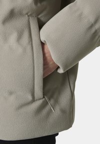 Lichtgrijze gewatteerde jacket met ribbelstructuur, grote zijzak en elastische manchetten. Ritsdetail op de zak.
