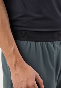 Detailný záber na ruku osoby, čierne sieťované tričko a sivé nohavice so širokým čiernym pásom s nenápadným tmavým nápisom.